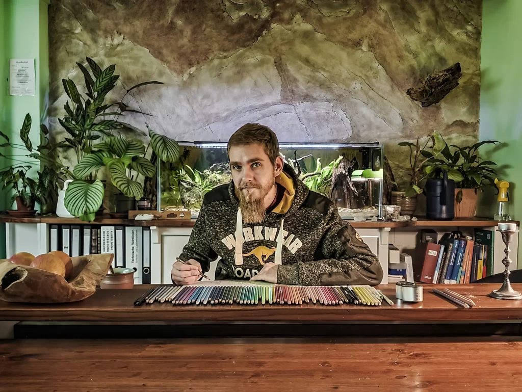 Künstler Kenny sitzt am Schreibtisch und arbeitet an einer Zeichnung mit Tiefe.